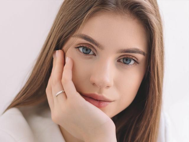 EricaDiaz has blue eyes, brown hair, and is touching her cheek with a hand wearing a silver ring, looking softly at the camera. Her lips are slightly parted.