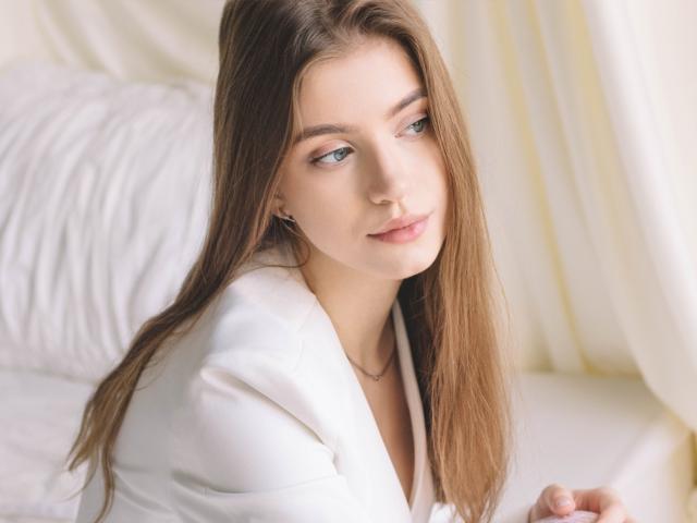 EricaDiaz has long brown hair, blue eyes, and a slight smile, wearing a white top, with a subtle necklace, sitting on a bed with white pillows.