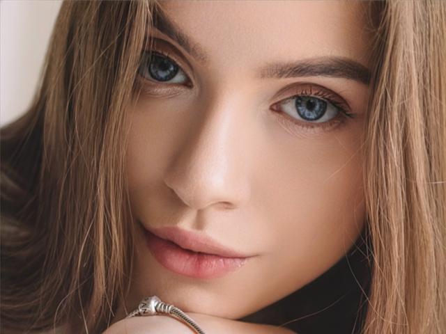 EricaDiaz's face, featuring blue eyes, full pink lips, and brown hair, with a silver ring on her finger resting on her chin. Her skin is smooth and fair.