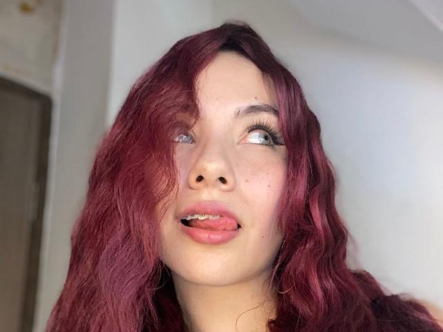ValeryHarllow has red wavy hair, light skin, blue eyes, and is sticking her tongue out slightly. Her long eyelashes are visible. She's looking to the right.
