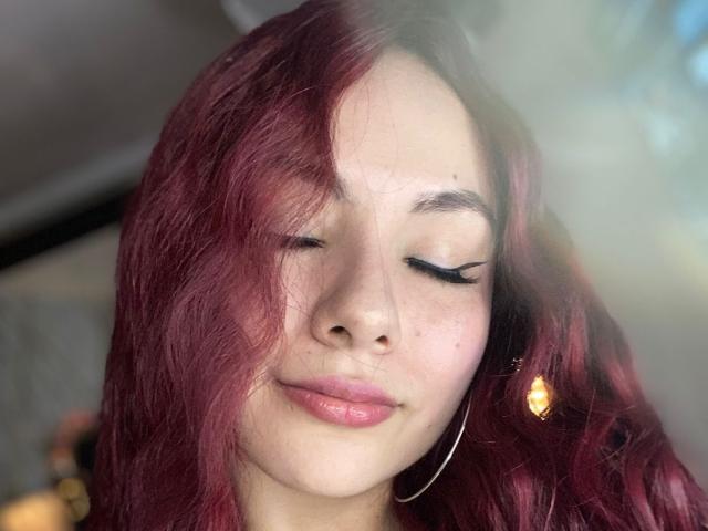 ValeryHarllow with red hair, closed eyes, and black eyeliner, wearing a gold earring and hoop, has a slight smile and pink lips, sunlight in the background.
