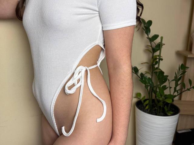 Symphoniia's white ribbed one-piece with side ties, showing her ass and part of her thigh. She's standing with a potted plant on the right.