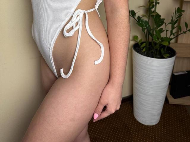 Symphoniia's white ribbed one-piece shows her bare ass and sideboob, tied with strings on the left. Her pink nail touches her thigh. Potted plant in the background.