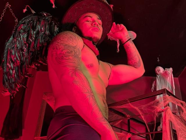 Muscular tattooed JeisonStark in a cowboy hat, red-lit, holding a rope, shirtless, wearing black pants, with a hanging bird and a wrapped-up figure in the background.