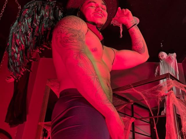 Muscular JeisonStark with tattoos, wearing a cowboy hat and dark pants, holding a toy skeleton, red lighting, and black wings in the background. His cock is not visible.