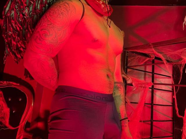 JeisonStark with red-tinted skin, tattooed arms, and blue-green dreadlocks, wearing tight Under Armour boxer briefs, standing in a red-lit room with spider web decoration.