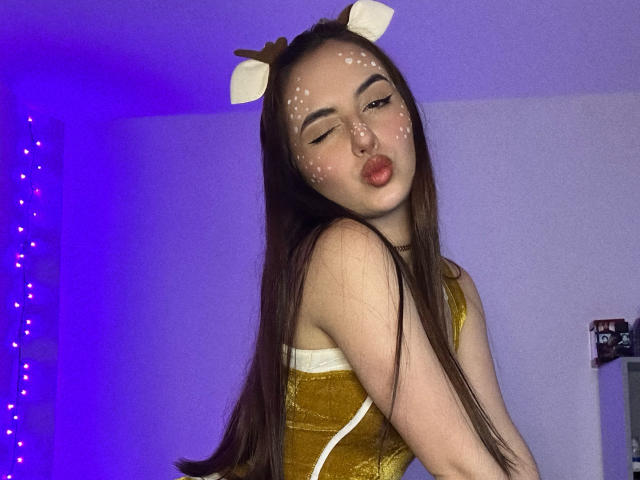 MiaHarperr with long brown hair, white deer antlers, and white face spots, pouting her red lips, wearing a yellow velvet dress, in a purple-lit room.
