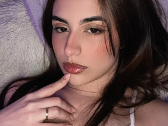 MiaHarperr has dark brown hair, brown eyes, and shiny pink lips. She's touching her chin with a finger, wearing a white top and a gold ring.