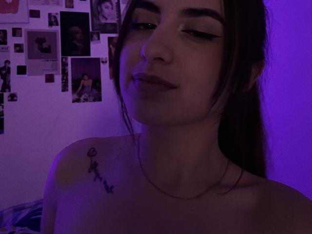 MiaHarperr ha i capelli castano scuro raccolti in una coda di cavallo, un trucco leggero e un piccolo tatuaggio sulla spalla sinistra. È senza top e illuminata da luci viola. Le sue natiche non sono visibili..
