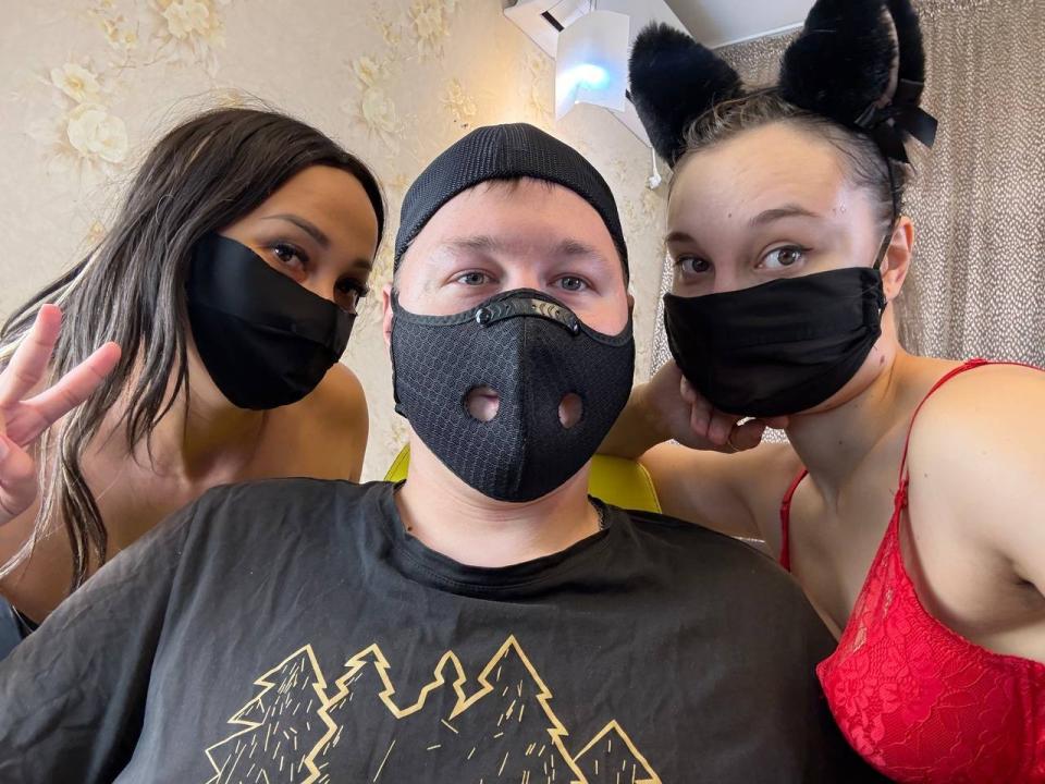 Three people, mask-covered faces, black beanie, black shirt with mountain graphic, red lace bra, black cat ears. SexLove69X in center, flanking two SexLove69X with hands up.