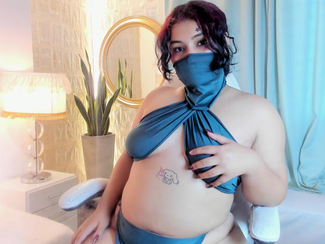 A photograph of AngelithaParker with curly dark hair and medium brown skin, wearing a teal halter top that covers her breasts but exposes her left nipple and a matching mask. She has a tattoo of two small hearts on her right side. Her left hand is covering her right breast, with dark red nails. She's sitting in a white chair, with a lamp, potted plant, and circular mirror in the background. The lighting is soft, with a blue and yellow glow.