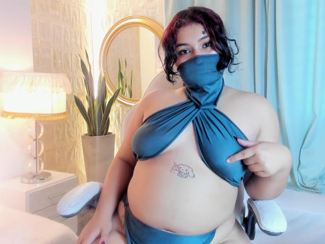 AngelithaParker, a plus-sized woman with curly black hair, wears a blue halter top and matching mask. She points at her chest, revealing a small tattoo of a fish below her left breast. She sits in a white chair with a potted plant and a lamp in the background. The image has soft lighting and a relaxed, intimate setting. Her skin is light brown.