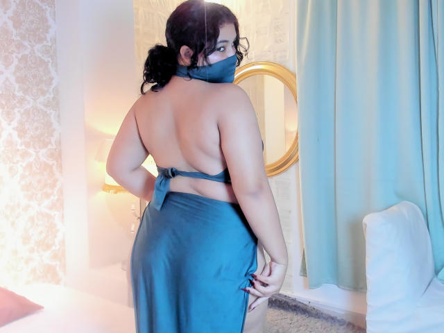 Angelitha Parker, con cabello negro rizado, vestida con una máscara azul y un vestido azul sin mangas, mostrando su trasero y espalda desnudos, está de pie en una habitación con un espejo dorado..