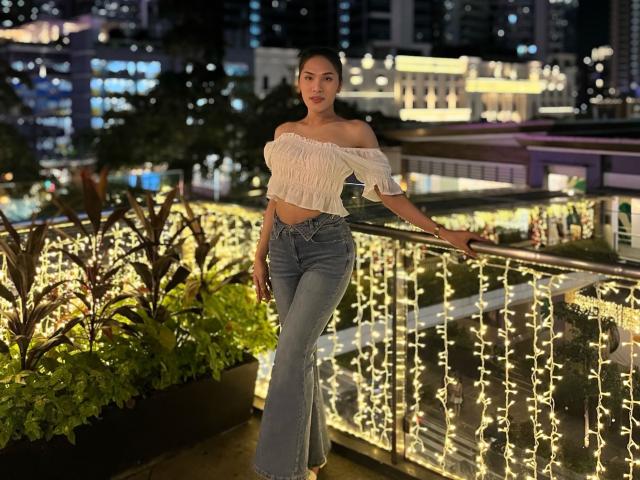 SofiaCumness, avec de petites seins, portant un haut blanc à une épaule et un jean bleu taille haute, appuyée contre un balcon éclairé de guirlandes la nuit. Ses cheveux noirs sont lâchés..