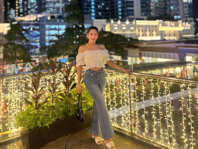 SofiaCumness with cute tits, off-shoulder white top, blue jeans, holding black purse, standing on balcony, string lights, city lights in background, wearing white sandals, night time.