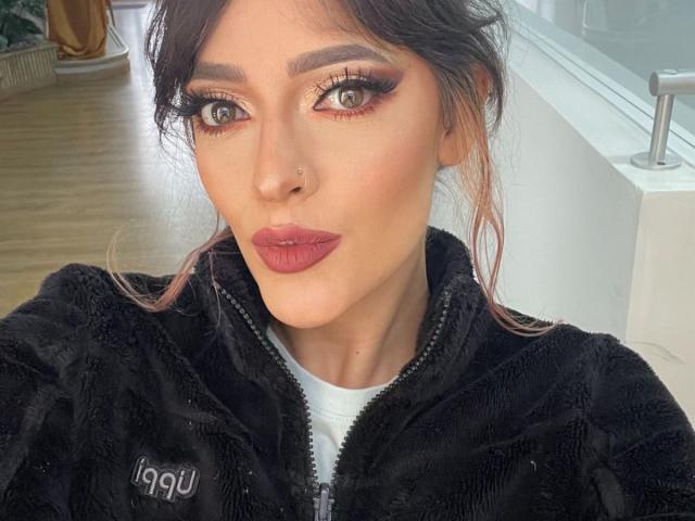 JaneRoa with pouty pink lips, green eyes, and dark makeup, wearing a black fuzzy zip-up, has pink-tinted hair, and a nose piercing, in a selfie.