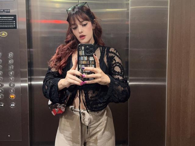 JaneRoa with red hair, black lace top, and white pants, taking a mirror selfie in an elevator. She's holding a phone with a grid pattern case, pink nails.