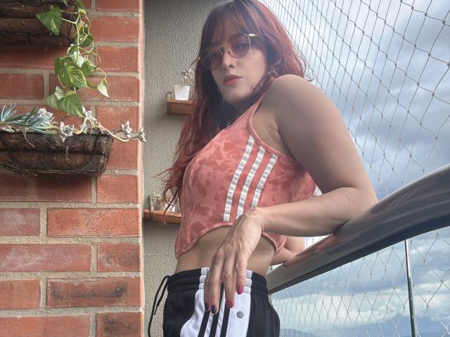 JaneRoa, con cabello rojo, gafas, un top deportivo rosa con estampado tie-dye y pantalones cortos negros y blancos, apoyada en una barandilla de metal, con las manos en las caderas, mirando hacia arriba. Sus pechos son pequeños..