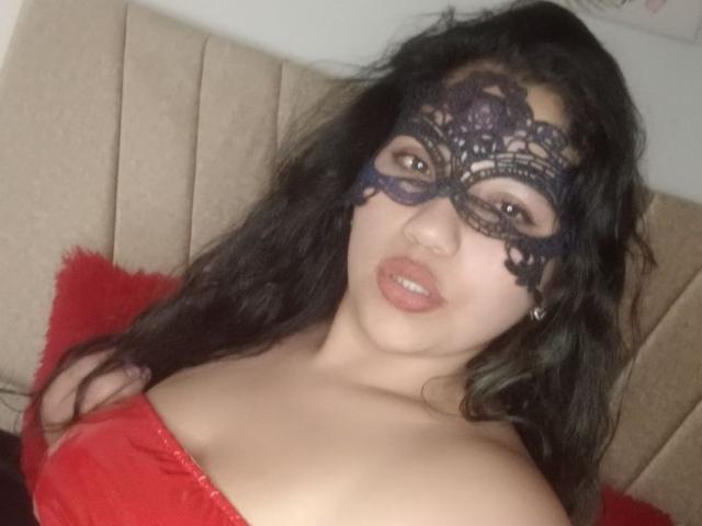 SallomeHotty, con cabello negro ondulado, vistiendo una máscara de encaje negro y un top rojo sin tirantes, mostrando sus grandes senos y labios rojos ligeramente entreabiertos, mirando a la cámara..
