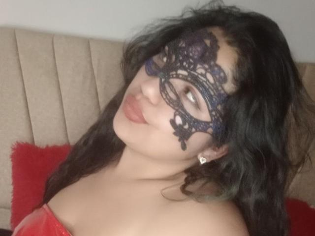 SallomeHotty met een donker kantmasker, golvend zwart haar en rode lippenstift, die lichtjes glimlacht en haar borsten laat zien, gekleed in een rode top zonder bandjes en met een kleine diamanten oorbelle..
