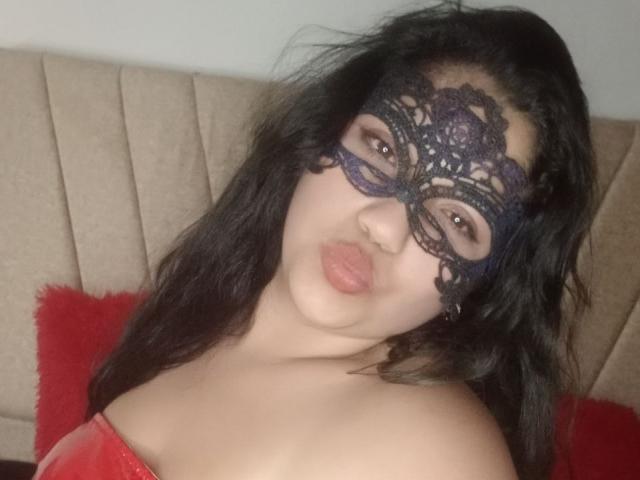 SallomeHotty with black lace mask, pouty lips, and dark wavy hair, wearing red off-shoulder top, showing her bare shoulders and upper chest, looking sexy.