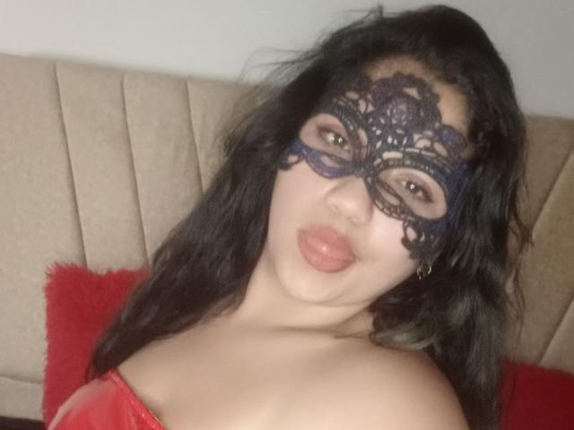 SallomeHotty with black lace mask, pouty pink lips, and wavy dark hair, wearing red strapless top, showing off her boobs and cute face.
