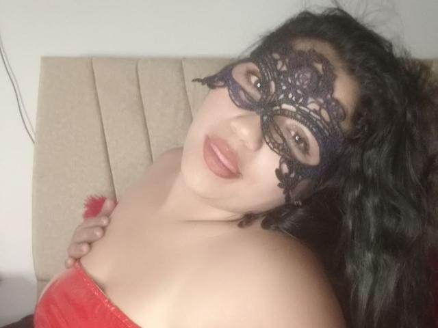 SallomeHotty with dark curly hair, wearing a black lace mask, red strapless top, and red fluffy accessory, smiling at the camera, showing her bare shoulders and tits.