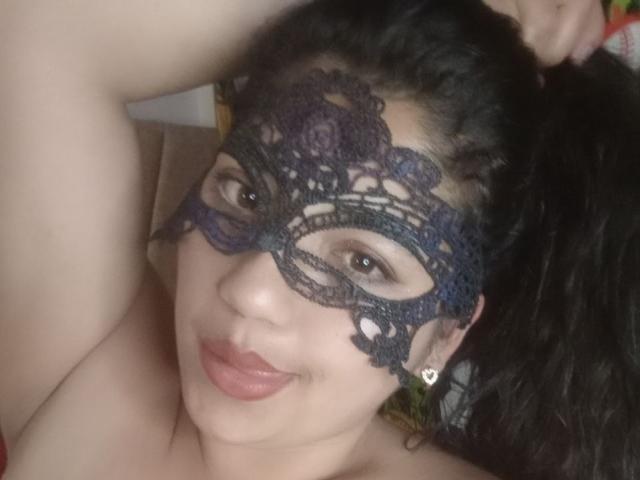 SallomeHotty with dark curly hair, wearing a black lace mask, smiling, showing her pouty lips, and holding her ponytail up. Her tits are partially visible.
