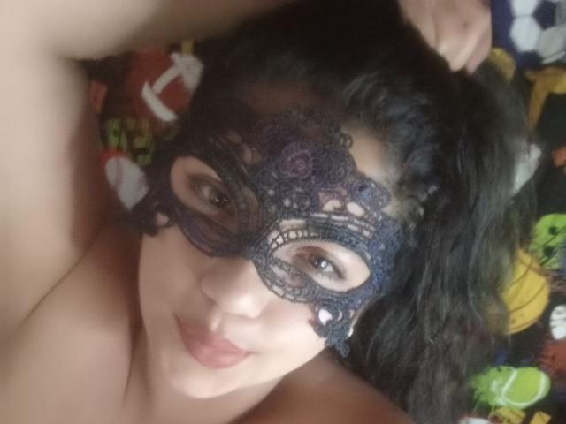 SallomeHotty with dark curly hair, wearing a black lace mask, lying down, topless, smiling softly, hand in hair, pale skin, brown eyes, and a playful, sexy expression.