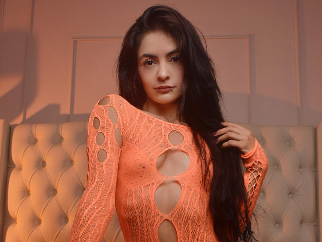 CandiceValo with long black hair, wearing a neon orange, cut-out, long-sleeve knit top, showing her small boobs and nipples through the holes. Standing in front of a tufted bed.
