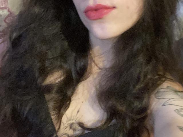DianaLiana with wavy black hair, red lipstick, cleavage, and a tattoo on her right shoulder. Boobs slightly visible. Skin is pale. Photo is a selfie.