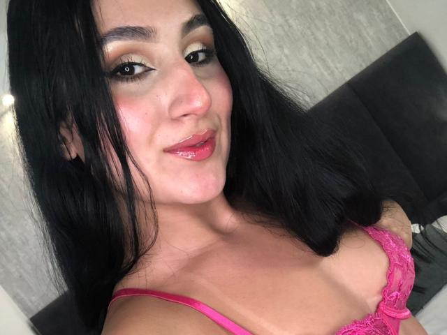 HiilaryDolce with black hair, pink lace bra, shiny pink lips, and slight smile, showing her tits and cute face. She's got a sexy, confident look.