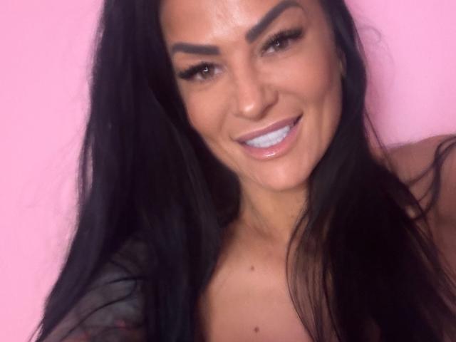 AndraD has black hair, thick eyebrows, blue teeth, and a shiny smile. She's smiling with her teeth showing, wearing a sheer top against a pink background.