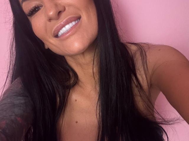 Close-up selfie of AndraD with a bright pink background. AndraD has long, straight black hair, a fair skin tone, and is smiling with white, slightly glossy teeth. Her right arm is partially visible, showing a tattoo with dark, intricate patterns. She has subtle, natural makeup with a light pink lip color. Her eyes are slightly upcast, giving a relaxed, friendly expression. The focus is on her face and upper chest. The image is sharp with clear details on her hair and tattoo. AndraD is looking slightly to the left of the camera.