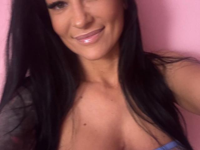 AndraD has long black hair, smiling with glossy pink lips, and visible cleavage in a blue top. She has a tan skin tone and a small mole on her chest.