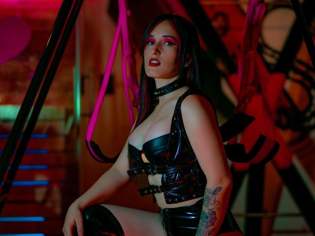 DominatrixEm with black leather bra, straps, and choker, showcasing her tits and tattooed arm, in a sexy, dark, neon-lit setting, with black thigh-highs and red lipstick.