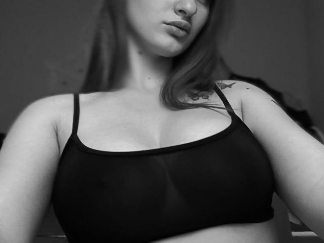 Reggiseno nero, seno di Millania, piccolo tatuaggio sulla spalla destra, parte del viso, labbra piene, lunghi capelli castani, scala di grigi, luce soffusa, parte superiore del corpo, ombra leggera, casual, sicura di sé..
