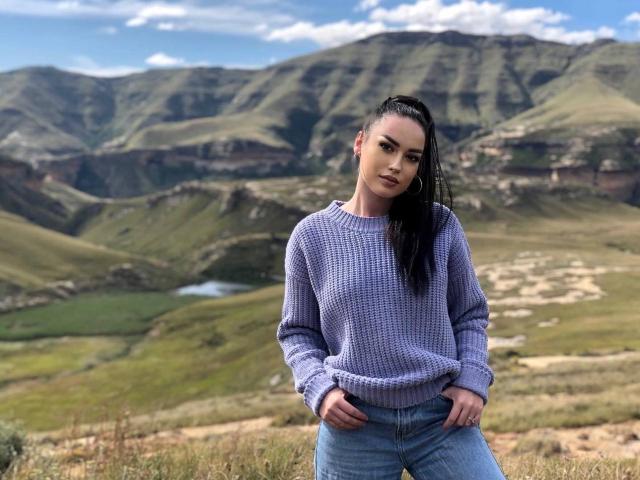 AnabelaBeauty with long black hair in a ponytail, wearing a purple knitted sweater and blue jeans, standing in a grassy mountain landscape, hands in pockets, looking at camera.