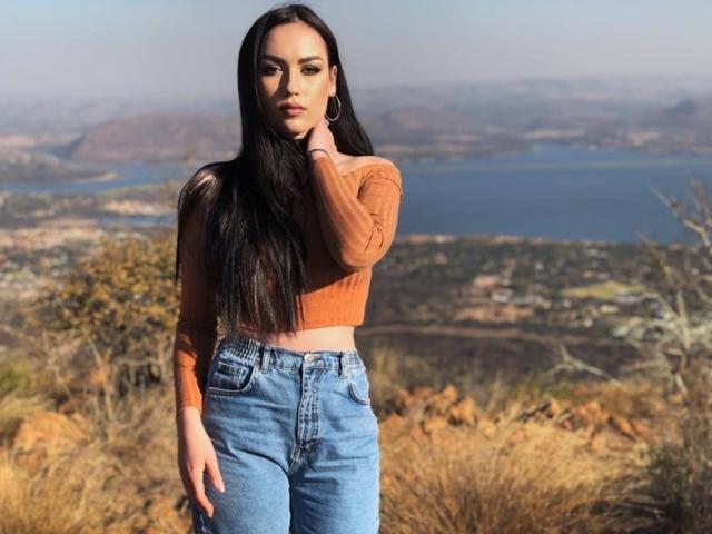 AnabelaBeauty with long black hair, orange crop top, and blue jeans, standing outdoors, hand touching her face, looking serious, natural light, clear sky, blue water in background.