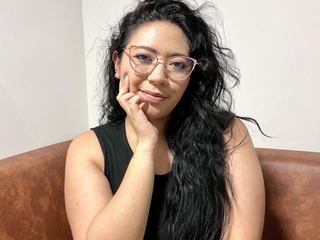 ErikaFox, com cabelos longos, pretos e desalinhados, usando óculos rosa e uma blusa preta sem mangas, apoiando o queixo na mão, sorrindo suavemente. Sofá de couro marrom ao fundo..