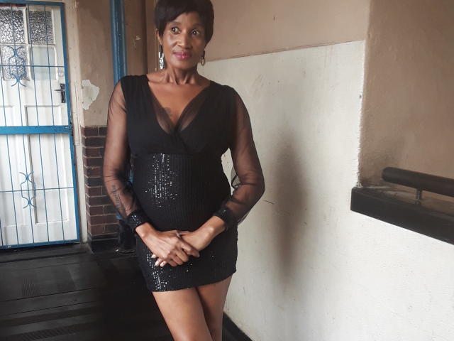 KinkyPussyLips, a dark-skinned woman with short black hair, wearing a black sheer dress with sequins, standing with hands together, wearing silver earrings, and a confident smile.