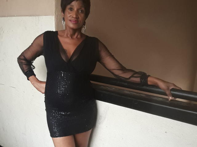KinkyPussyLips in a black sheer dress with sparkly sequins, standing with one arm on a railing, wearing earrings, smiling, showing her ass and small tits.