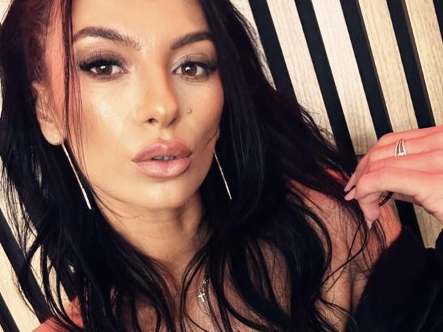 KinkyAmelie hat langes, schwarzes Haar, braune Augen und volle, rosafarbene Lippen. Sie trägt große goldene Ohrringe und ein schwarzes Oberteil, und sie hat einen Ring an ihrer rechten Hand..