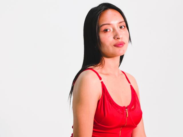 NicolleGlory, with long black hair, wearing a red dress, has small tits, a cute face, and red lipstick, standing against a white background.