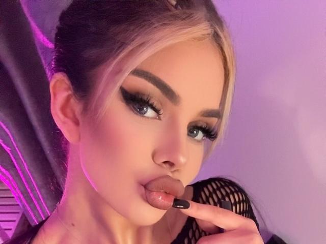 IvyRaven with blonde hair, thick eyebrows, and long eyelashes, sucking her black-nailbed finger, looking seductive with pink-purple lighting. She has a cute face and is wearing fishnet.