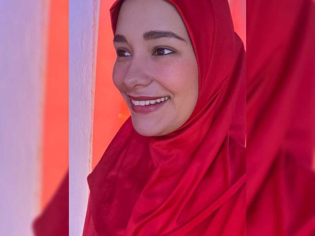 ShanaJamil with red hijab, smiling, showing white teeth, brown eyes, and fair skin, her face slightly turned, red fabric covering her head and body, soft light on her face.
