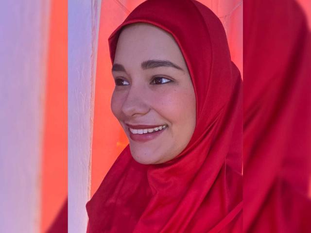 ShanaJamil with red hijab, smiling, showing white teeth, looking to the side, wearing red, her ass slightly visible in the background, bright red light behind her.