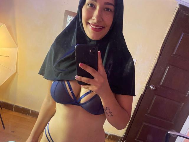 ShanaJamil with a black hijab, smiling, wearing a blue bra with strappy details, showing her tits, and a matching blue thong, holding a phone in a mirror selfie.