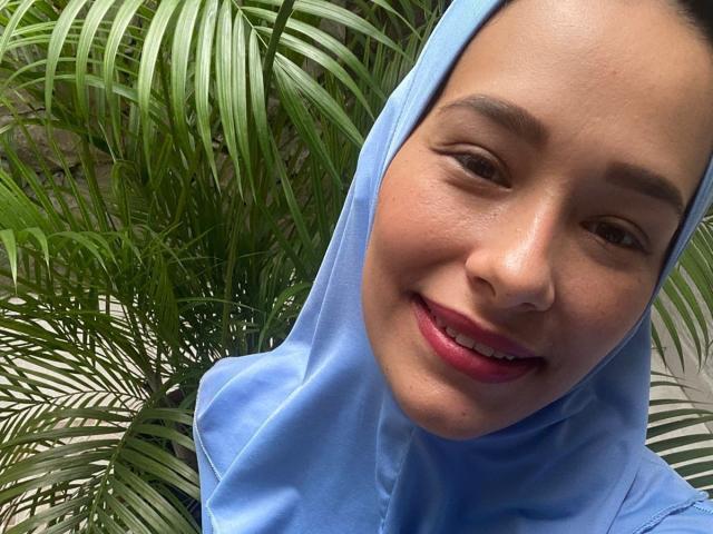 ShanaJamil with light brown skin, smiling, wearing a blue hijab, green palm leaves in the background, slightly wavy dark hair, pink lipstick, and a cute, relaxed expression.