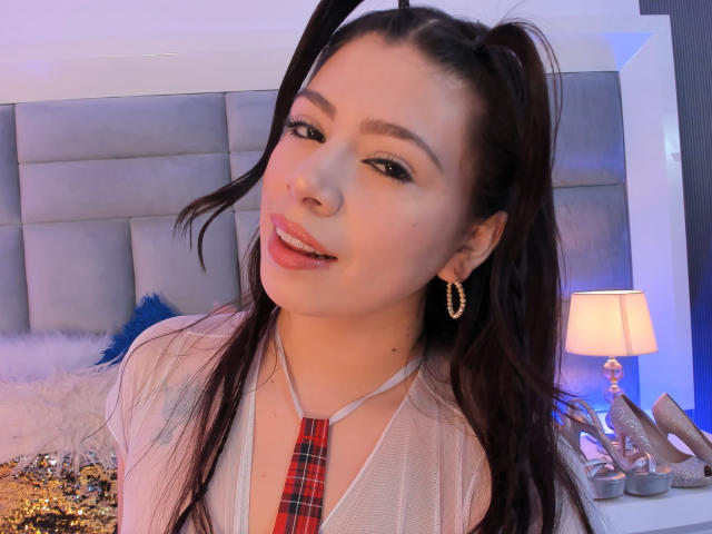 LindaMancini with long dark hair in pigtails, wearing a sheer top with a red plaid tie, gold hoop earrings, smiling, bed with fluffy pillow and lamp in background.